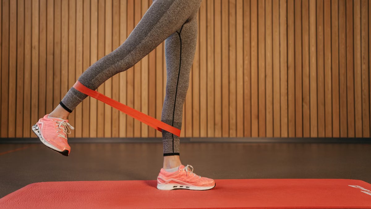 A woman performing leg exercises using a red resistance band on a yoga mat indoors.